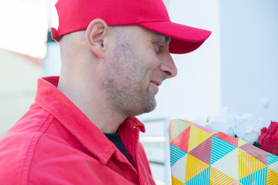A man wearing a red cap and red jacket is holding a colorful gift bag with a geometric pattern of yellow, blue, red, and white triangles, containing white and red flowers. The background is slightly b