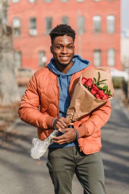A young man with dark curly hair and a cheerful expression is standing outdoors, holding a bouquet of fresh red roses wrapped in brown kraft paper. He is dressed in a bright orange puffer jacket layer