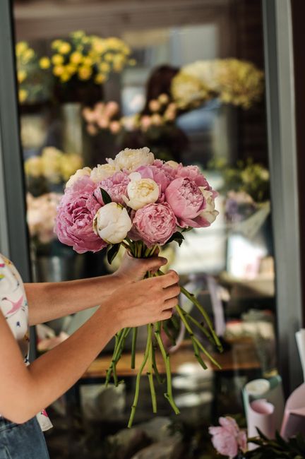 A person's hands holding a bouquet of fresh pink and white peonies, surrounded by green stems, with a glass florist display case in the background showcasing various floral arrangements and potted pla