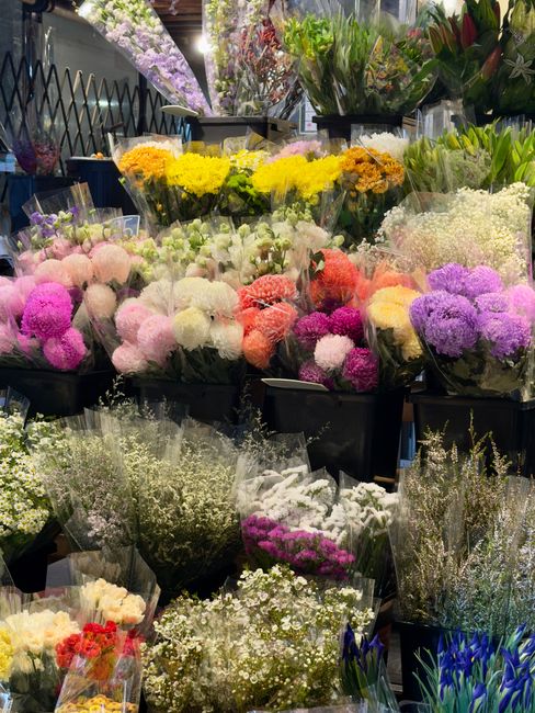 A vibrant display of fresh floral arrangements at a flower shop, featuring clusters of chrysanthemums, carnations, and baby's breath in shades of pink, purple, yellow, white, and peach. The bouquets a