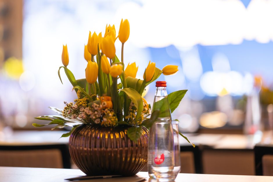 A floral arrangement featuring a bouquet of bright yellow tulips with closed and partially open petals, accented by small pink and white filler flowers and lush green leaves, is artfully placed in a r