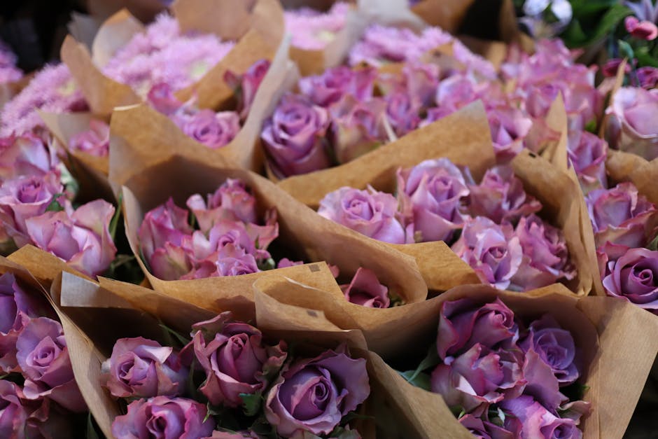 A collection of fresh pink and lavender roses, each with delicate, velvety petals, arranged in small bouquets wrapped in brown kraft paper. The roses are densely packed, with visible green stems and b