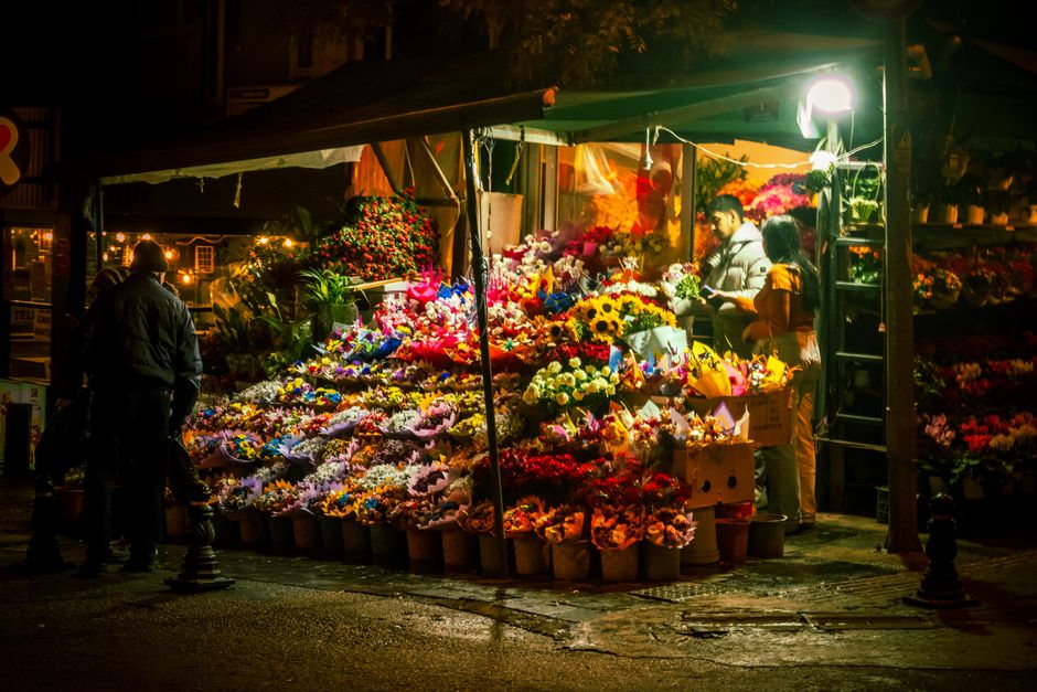 A night scene at a flower stall near Blackfriars Station, illuminated by bright artificial lighting. The stall displays a wide variety of fresh flowers, including vibrant sunflowers, white roses, pink
