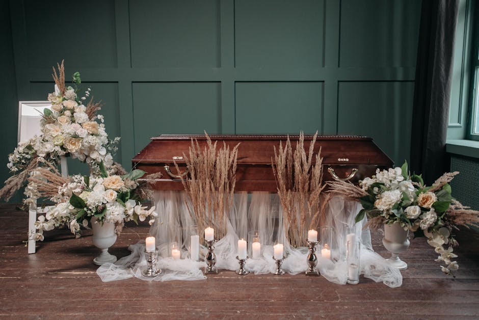 A floral arrangement featuring predominantly white and soft pastel flowers, including roses and hydrangeas, displayed in elegant white vases on either side of a wooden casket. The arrangement is accen