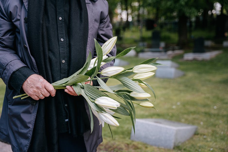 A person dressed in a dark jacket and black shirt is holding a bouquet of white lilies with green leaves, arranged with the flowers facing outward. The bouquet is wrapped in clear plastic, and the per