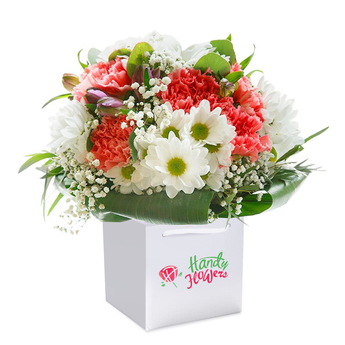 Resting upright in a sharp, white square gift bag, this bouquet draws the eye with its vibrant colour palette and expert craftsmanship-a genuine reflection of the artistic spirit thriving near Blackfriars Road. Waves of coral and vibr ant red carnations, each one forming a soft mound of ruffled petals, bring warmth and vitality, echoing the lively ambiance of nearby Southbank cafes. Among them, clusters of pristine white daisies with striking yellow-green hearts provide a dance of freshness and light. Delicate sprays of baby's breath intertwine throughout, their tiny blossoms lending an ethereal softness and visual lift, much like the clouds rolling over the London skyline. On the left edge, closed purple-tinged buds introduce depth-a visual whisper of the unfolding seasons, reminiscent of spring morning walks through Christ Church Garden. The entire floral display is encircled by robust, dark green foliage, glossy and strong, giving the bouquet balance and a sense of contemporary elegance. The gift bag itself, topped with sturdy white ribbon handles, hints at thoughtful delivery by Blackfriars Flowers, whether destined for an anniversary at home or a thoughtful gift for a colleague on Blackfriars Road. Crisp professional lighting against a clear white background ensures every stem and shade stands out in perfect harmony. This bouquet, both sophisticated and uplifting, celebrates the enduring link between nature's beauty and the heart of urban London.