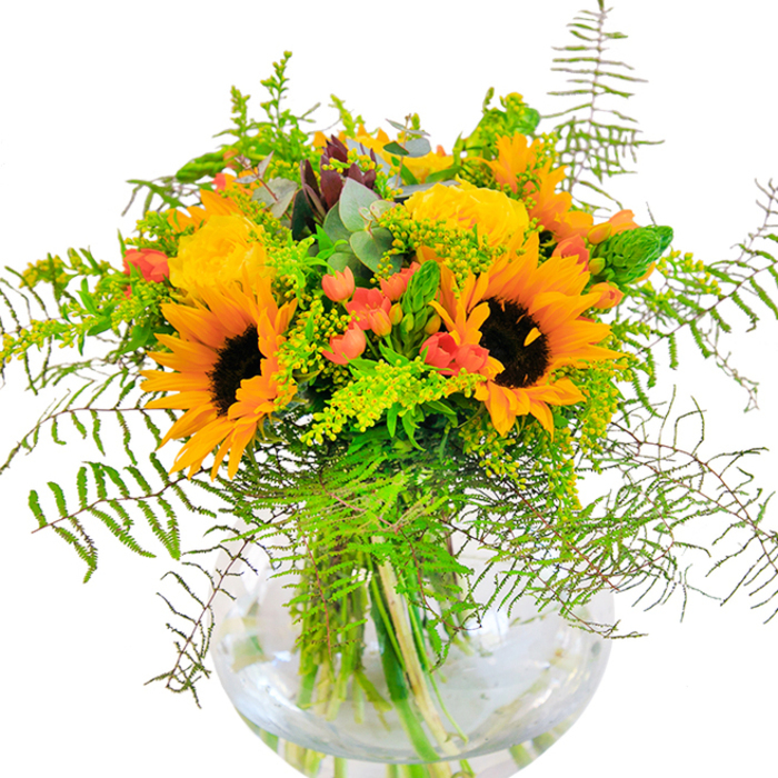 One glance at this sumptuous bouquet conjures a sense of sun-drenched optimism, firmly grounded in the heart of Blackfriars. The arrangement, beautifully suited for loft apartments overlooking the Thames or neighbourhood cafés, is centred on magnificent sunflowers-vivid yellow-orange petals spiralling around chocolate brown centres, each one turned as if to follow the arc of city light. Intermingled, golden yellow garden roses bloom in rich, sculpted layers, their texture velvety and inviting, bringing to mind the refined elegance of historic Blackfriars passageways. Between the blooms, clusters of bright red hypericum berries glisten brightly, their playful hues offering a charming counterpoint to the dominant yellows. A profusion of deep green foliage, enhanced with feathery ferns, cascades gracefully, recalling the green enclaves tucked between neighbourhood streets. Delicate sprigs of solidago add tiny yellow sparks of colour, while slender green flower spikes and bluish eucalyptus leaves provide freshness and textural complexity. Set within a crystal-clear, round glass vase, the arrangement's stems are crisply displayed, anchoring the liveliness above with a sense of transparency and order. The bouquet is ideal for marking joyful life events-a birthday toast, a riverside gathering, or simply brightening a grey London day. Its soft, sweet aroma hints at flower stalls in local markets and leafy river walks, embodying a perfect blend of rustic charm and urban sophistication from Blackfriars Flowers.
