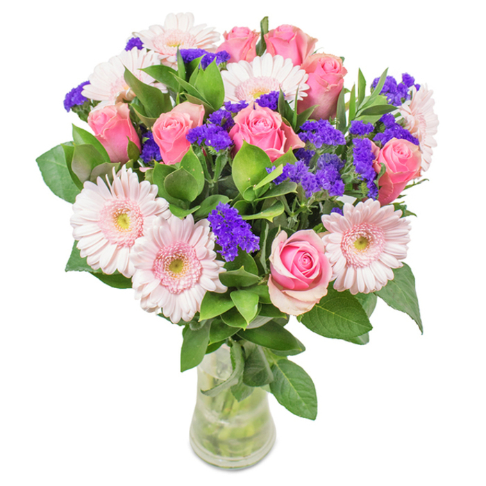 This eye-catching bouquet, crafted by Blackfriars Flowers and set against a clean white backdrop, showcases a harmonious blend of approximately seven to nine soft and mid-pink roses, their petals tightly furled or opening in classic spiral elegance reminiscent of blooms glimpsed through local garden gates. These roses are complimented by a gathering of five to seven pale pink Gerbera daisies, their large, round heads framed by feathery petals and almost-white centres, echoing the airy vibes of nearby riverside parks. Bright, vivid clusters of purple statice weave throughout, lending both rich texture and a jewel-toned accent that lifts the entire composition. Lush, deep green foliage forms a generous base, enhancing the display's freshness and vibrancy, while a crystal-clear vase allows the stems and sparkling water to anchor the arrangement in natural simplicity. Designed to delight for birthdays, anniversaries, or as a heartfelt gesture, this arrangement brings the refined beauty and community warmth of Blackfriars to home or office, infusing each space with joy and affectionate celebration.